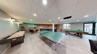 Located in Calgary, AB, this generously sized recreation room offers a dynamic, open-concept layout designed for leisure and social gatherings. The space features light-toned tile flooring throughout, a drop-panel ceiling with recessed pot lighting, and a combination of exposed brick, vertical wood slat paneling, and smooth painted walls that together create a modern yet inviting atmosphere.

The gaming area forms the heart of the room, anchored by a full-size Brunswick ping-pong table and a pool table with green felt. A foosball table is tucked into a dedicated corner alongside a wall-mounted cue rack, offering additional entertainment options. A glowing "Game On" neon sign adds a vibrant, energetic accent to the space.

A wet bar with dark cabinetry, bar seating, and a brick backsplash provides a social hub for refreshments. The lounge area features a round poker table surrounded by tan leather armchairs set atop a patterned area rug, offering a relaxed setting for card games. Framed landscape artwork decorates the walls, complemented by a large scenic print in the ping-pong zone.

Natural light filters in through multiple windows, and a glass-panel exterior door provides direct outdoor access. Lush potted plants are placed throughout, adding a fresh, welcoming touch to this well-equipped community recreation space.