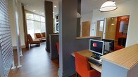 This single-floor space presents a well-organized, modern interior designed to balance functionality with a welcoming ambiance. The space is characterized by bold charcoal-gray structural columns that define distinct zones without fully enclosing them, creating an open yet structured layout. Rich hardwood flooring runs throughout, adding warmth to the otherwise contemporary aesthetic.

A workstation area features wooden countertops paired with vibrant orange seating, providing a practical and visually striking setup equipped with desktop computers. The reception or service counter nearby echoes the same wood-tone finish, maintaining a cohesive design language throughout the space.

Large windows along one wall flood the interior with natural light, offering views of greenery outside and enhancing the overall brightness of the space. The lounge area, visible in the background, features patterned upholstered seating in warm earth tones, offering a comfortable waiting or relaxation zone. Recessed lighting and pendant fixtures provide layered illumination throughout. The combination of dark columns, wood surfaces, and bold accent colors creates a polished, professional atmosphere suitable for a business, leasing office, or community hub.