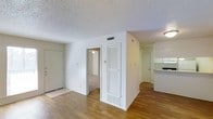 This single-floor space offers a clean, functional layout designed for comfortable everyday living. The main living area is anchored by warm hardwood-style flooring that extends throughout much of the unit, creating a cohesive and inviting atmosphere. A textured popcorn ceiling adds a classic touch overhead, while HVAC vents are thoughtfully integrated throughout for year-round climate control.

The open layout allows for a natural flow between the living area and the adjacent kitchen, which is visible from the main space and features white cabinetry and light countertops. A convenient pass-through or bar-style counter connects the kitchen to the living area, enhancing the sense of openness.

A set of louvered bifold doors provides access to a utility or storage closet, adding practical storage solutions within easy reach of the main living space. A doorway leads to a carpeted room, likely a bedroom, offering a softer, quieter retreat from the rest of the unit. Additional doors suggest further rooms or exterior access points, contributing to the overall versatility of the floor plan.

The bright white walls and neutral palette throughout keep the space feeling light and airy, while the straightforward design makes it easy to personalize with furnishings and décor.