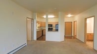 This single-floor residence offers a practical and well-organized open-concept layout that seamlessly connects the living and kitchen areas. The spacious living room is finished with wall-to-wall carpet, providing a comfortable and inviting atmosphere, while a baseboard heating unit runs along the wall for efficient climate control.

The kitchen is defined by light oak cabinetry that extends to the upper walls, offering generous storage throughout. Black appliances, including a range, microwave, and refrigerator, provide a clean contrast to the warm cabinetry tones. A countertop peninsula with a raised breakfast bar serves as a natural divider between the kitchen and the living space, enhancing both functionality and flow. The kitchen floor transitions to a hard surface, distinguishing it from the carpeted living area.

Structural columns frame the kitchen opening, adding architectural character to the space. A hallway visible from the living area leads to additional rooms, including what appears to be a bathroom with cabinetry. A solid wood interior door provides access to further areas of the unit. The neutral color palette throughout — warm beige walls, light wood tones, and subtle flooring — creates a cohesive and understated aesthetic across the entire space.
