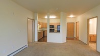This single-floor residence offers a practical and well-organized open-concept layout that seamlessly connects the living and kitchen areas. The spacious living room is finished with wall-to-wall carpet, providing a comfortable and inviting atmosphere, while a baseboard heating unit runs along the wall for efficient climate control.

The kitchen is defined by light oak cabinetry that extends to the upper walls, offering generous storage throughout. Black appliances, including a range, microwave, and refrigerator, provide a clean contrast to the warm cabinetry tones. A countertop peninsula with a raised breakfast bar serves as a natural divider between the kitchen and the living space, enhancing both functionality and flow. The kitchen floor transitions to a hard surface, distinguishing it from the carpeted living area.

Structural columns frame the kitchen opening, adding architectural character to the space. A hallway visible from the living area leads to additional rooms, including what appears to be a bathroom with cabinetry. A solid wood interior door provides access to further areas of the unit. The neutral color palette throughout — warm beige walls, light wood tones, and subtle flooring — creates a cohesive and understated aesthetic across the entire space.