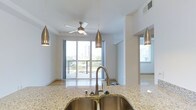 This single-floor residence offers a thoughtfully designed open-concept layout that seamlessly connects the kitchen and living area. The kitchen is a standout feature, showcasing rich dark cabinetry that contrasts beautifully with light granite countertops. A central island with a double undermount stainless steel sink serves as a functional and stylish focal point, while sleek pendant lights with a brushed gold finish hang overhead, adding a contemporary touch. Stainless steel appliances, including a refrigerator and range, complete the kitchen's modern aesthetic. Warm hardwood flooring flows throughout, enhancing the bright, airy feel of the space. The living area is bathed in natural light from a large sliding glass door that opens to a balcony with expansive city views, offering a striking urban backdrop. A ceiling fan keeps the space comfortable year-round. Elevated ceilings with a tray detail in the kitchen add architectural interest and a sense of volume. A full bathroom, visible from the kitchen corridor, features a tub and shower combination with clean, neutral finishes. Overall, this residence combines functional design with modern finishes to create a polished and inviting living environment.