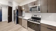 This contemporary 2-bedroom, 1-bathroom unit offers a well-designed, single-floor layout with modern finishes throughout. The open-concept kitchen and living area serves as the heart of the unit, featuring light hardwood-style flooring, recessed lighting, and a clean, neutral color palette. The kitchen is outfitted with dark wood cabinetry, white quartz countertops, a classic subway tile backsplash, and a full suite of stainless steel appliances including a refrigerator, electric range, microwave, and dishwasher. The living area flows seamlessly toward floor-to-ceiling sliding glass doors that open to a private balcony, offering views of the surrounding urban landscape. Both bedrooms are carpeted with a multi-tone gray patterned carpet and feature large windows that bring in natural light and city views. The primary bedroom is a comfortable, well-proportioned space. The shared bathroom is crisp and modern, highlighted by an all-white walk-in shower enclosure with a frameless glass door, chrome fixtures, and a built-in niche. A dedicated closet provides additional storage. Throughout the unit, clean lines, recessed lighting, and a cohesive neutral palette create a bright, functional, and modern living environment.