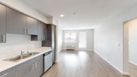 This single-floor unit offers a clean, contemporary design with an open-concept layout that seamlessly connects the kitchen and living area. The kitchen is well-appointed with sleek grey flat-front cabinetry extending to the ceiling for ample storage, white quartz countertops, stainless steel appliances including a refrigerator, electric range, and dishwasher, and a stainless steel sink. Light wood-look flooring runs throughout the main living areas, enhancing the bright and airy atmosphere created by recessed lighting and a large window that invites natural light into the living space. A door off the living area provides additional access and light. A hallway leads to the remaining areas of the unit, including a full bathroom outfitted with grey cabinetry, a vanity with sink, a bathtub and shower combo, and wood-look flooring. A spacious walk-in closet features built-in shelving and hanging rods, and is finished with plush carpet flooring, offering generous storage capacity. The overall ambiance of the unit is modern and well-maintained, with a neutral color palette of whites and greys that provides a versatile backdrop throughout.