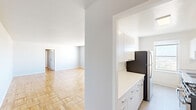 This single-floor unit presents a clean, functional layout across multiple rooms, unified by a consistent neutral color palette of white and light grey. Warm-toned parquet wood flooring runs throughout the living areas and bedrooms, adding visual texture and character to the space.

The kitchen is divided into two distinct areas. The first features sleek white cabinetry extending to the ceiling, a white countertop, and a stainless steel refrigerator alongside a dark built-in appliance. The second kitchen area is brighter, benefiting from a large window that brings in natural light and frames an elevated outdoor view of surrounding greenery. This section includes a stainless steel double-basin sink, additional white upper cabinets, and a stainless countertop.

The living area is generously proportioned, with smooth white ceilings and parquet flooring that extends seamlessly into the connecting hallway and bedrooms. The bedrooms are well-sized, each featuring the same parquet flooring and neutral walls. At least one bedroom includes a sliding closet with a built-in rod, while another opens directly to a bathroom equipped with a pedestal sink and toilet. The overall ambiance is bright, minimal, and move-in ready.