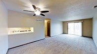 This single-floor residence offers a functional and comfortable layout with a welcoming open-concept living area at its heart. The main living space features plush wall-to-wall carpeting underfoot and a textured ceiling overhead, creating a cozy and practical environment. A ceiling fan with an integrated light fixture provides both airflow and ambient lighting, ensuring year-round comfort. Natural light fills the room through a sliding glass door, which opens to an outdoor area and frames views of the surrounding landscape. A convenient pass-through counter connects the living area to the kitchen, allowing for easy interaction between spaces. The kitchen area is visible through this opening and includes a sink and countertop workspace. A hallway off the main living area leads to additional rooms, with a dark-toned door providing a subtle design accent. HVAC ventilation is thoughtfully integrated into the upper wall, ensuring efficient climate control throughout. The overall ambiance is clean, open, and move-in ready, with a neutral color palette that offers a versatile backdrop for a variety of interior styles.