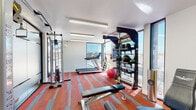 This fitness center offers a comprehensive workout environment designed to accommodate a wide range of exercise routines. The space is laid out across a single floor and is divided into distinct training zones, each thoughtfully arranged for functionality and ease of use.

The flooring throughout features a bold striped carpet in shades of red and gray, lending the space a vibrant, energetic atmosphere. Bright fluorescent lighting runs along the white ceilings, ensuring consistent, even illumination across all areas.

Natural light is a standout feature, with large floor-to-ceiling windows and glass panels lining the perimeter. These expansive windows frame outdoor views and allow generous amounts of daylight to fill the interior, creating an open and airy feel.

The cardio zone is positioned near the windowed walls and includes a Spirit treadmill, offering users an outdoor view while exercising. The strength training area is well-appointed with a multi-functional cable machine, a lat pulldown station, adjustable weight benches, and a dedicated training zone rack stocked with medicine balls and kettlebells. TRX suspension straps are also available for functional training.

A separate section houses a dumbbell rack with a full range of free weights, providing space for targeted strength work. Wall-mounted mirrors and a split-system air conditioning unit contribute to a comfortable and practical training environment overall.