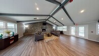 This single-floor space is defined by its striking architectural character and inviting atmosphere. The main lounge area commands immediate attention with its soaring vaulted ceiling adorned with bold, dark-painted exposed beams that create a strong visual framework overhead. Anchoring the room is a dramatic floor-to-ceiling natural stone fireplace wall, a standout feature that lends a rustic yet refined character to the space. A wall-mounted flat-screen television sits above the fireplace's slate mantel, while comfortable gray sofas arranged around a wood coffee table and a vibrant area rug complete the seating area. Warm hardwood flooring flows seamlessly throughout the entire space, enhancing the sense of continuity and openness. Large windows allow generous natural light to filter in, connecting the interior to the surrounding outdoors. An adjacent area offers bar-height tables with stools alongside a service counter, making it well-suited for gatherings and events. Colorful wall art adds contemporary accents, while a decorative console with a round mirror provides an elegant transitional element. Additional exit doors open directly to the exterior, further enhancing accessibility and airflow throughout this well-appointed, versatile space.