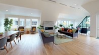 This single-floor space showcases a thoughtfully designed open-plan layout that seamlessly connects multiple living and dining areas beneath dramatically angled ceilings. Wide-plank hardwood flooring flows consistently throughout, lending warmth and cohesion to the entire space.

The main living area is generously proportioned, anchored by a floor-to-ceiling stacked stone fireplace that serves as a striking focal point. Multiple seating arrangements — including plush sofas and accent armchairs — define distinct zones within the open layout. Large casement windows on multiple sides of the space flood the interior with natural light and offer views of the surrounding neighborhood rooftops.

A dedicated lounge nook positioned near oversized windows provides an intimate seating area, ideal for reading or conversation. Adjacent to the main living zone, a dining area features a substantial wood table paired with modern chairs, alongside a wall-mounted display and a built-in wooden credenza with cabinet storage.

Throughout the space, lush indoor plants add organic texture and vitality, complementing the clean white walls and the warm tones of the hardwood floors. The overall ambiance is modern yet inviting, balancing architectural interest with a relaxed, well-curated aesthetic.