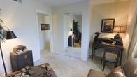 This single-story space includes a bedroom that serves as a functional and comfortable living area. The room is finished with wall-to-wall carpet in a neutral beige tone, contributing to a warm and quiet atmosphere. A standout feature is the full-length mirrored sliding closet, which visually expands the room by reflecting the surrounding space and adding depth. The mirrors also allow natural light to bounce throughout the room, enhancing the overall brightness.

To the right of the closet, a dark-wood writing desk and chair occupy a compact corner, accompanied by framed wall art that adds a decorative touch. The walls are painted in a soft, neutral tone that complements the light carpeting and contributes to the room's understated, clean aesthetic.

An open doorway leads to an adjacent room, suggesting a connected layout that promotes easy flow between spaces. The adjoining room, visible through the doorway, shares similar neutral finishes and carpeting, maintaining a cohesive design throughout. Overall, the space presents a simple, functional layout with classic residential finishes suited for everyday living.