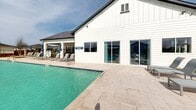 Located at 1551 Beltmill Pkwy in Saginaw, Texas, this community swimming pool facility offers a well-appointed outdoor retreat designed for relaxation and recreation. The centerpiece is a generously sized swimming pool with crystal-clear turquoise water, ranging in depth from the shallow end to deeper sections, with depth markers clearly visible along the pool's edge.

Surrounding the pool is a broad, stone-paved sun deck that provides ample space for lounging. Rows of modern sling-style chaise lounges with side tables line the pool deck, offering plenty of seating for residents. Patio umbrellas are positioned throughout for shade. A pergola structure with a wood-beam design anchors one end of the deck area, providing a covered gathering spot with additional seating.

Adjacent to the pool, a striking white board-and-batten clubhouse building features large sliding glass doors that open directly onto the pool deck, creating a seamless indoor-outdoor connection. Stone pillar accents and decorative fencing frame the perimeter of the facility, maintaining an upscale aesthetic while ensuring safety.

The surrounding landscaping includes mulched planting beds, decorative river rock borders, and young trees that will provide additional shade and greenery as they mature. The facility is enclosed by a sleek black metal fence, offering clear views of the surrounding residential neighborhood under the open Texas sky.