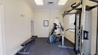 This single-floor fitness center offers a comprehensive workout environment designed for both cardio and strength training. The main exercise area features durable rubber-speckled flooring that provides cushioning and traction, complemented by warm, cream-colored walls adorned with crown molding for a refined aesthetic. Natural light streams in through multiple windows fitted with white plantation shutters, as well as a classic French-style glass-paned exterior door that opens to the outdoors. The space is outfitted with a multi-function cable machine for full-body strength training, an elliptical trainer, two stationary upright bikes, a treadmill, and an adjustable weight bench, providing a well-rounded selection of equipment. A wall-mounted flat-screen television is positioned for easy viewing during workouts. Recessed panel ceiling lights ensure consistent, even illumination throughout. An adjacent room with tile flooring and its own wall-mounted television provides a secondary space that can serve as a stretching, yoga, or cool-down area. French doors throughout create a seamless flow between areas. The overall ambiance is clean, bright, and functional, offering a welcoming environment for a complete fitness routine.