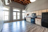 This single-floor unit showcases an industrial-loft aesthetic defined by its most striking feature: exposed wooden ceiling beams paired with visible ductwork and pendant lighting suspended from above. The open main living area is generously proportioned for its footprint, benefiting from soaring ceiling heights that amplify the sense of space. Large, arched windows flood the interior with natural light while framing urban cityscape views, contributing to a bright and airy atmosphere throughout.

The main living space incorporates an open kitchen along one wall, equipped with cream cabinetry, laminate countertops, and black appliances including a range, microwave, and refrigerator. A large sliding barn door serves as a architectural divider within the main space, adding a rustic contrast to the industrial elements overhead. Light-toned wood-style flooring runs continuously throughout, unifying each area.

A hallway connects the main living space to the remainder of the unit, featuring an arched entryway and additional pendant lighting beneath the exposed beam ceiling. The full bathroom includes a single-sink vanity with wood cabinetry, a tub-shower combination, and matching wood-style flooring, offering a clean and functional design consistent with the overall aesthetic of the space.