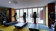 This single-floor fitness center is designed with both functionality and comfort in mind. The space is anchored by a row of modern treadmills equipped with individual display screens, positioned to take full advantage of the expansive floor-to-ceiling glass sliding doors that line the far wall. These large glass doors flood the room with abundant natural light and offer unobstructed views of an outdoor patio area, complete with seating and greenery, creating a seamless connection between the interior workout space and the outdoors. The clean, contemporary aesthetic is reinforced by recessed ceiling lighting, smooth white walls, and a warm wood-paneled accent wall that adds texture and visual interest. The flooring features a combination of gray and yellow tones, providing a practical yet visually dynamic surface suited for active use. The overall ambiance is bright, open, and inviting, making it a well-equipped and thoughtfully designed environment for fitness activities.