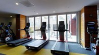 This single-floor fitness center is designed with both functionality and comfort in mind. The space is anchored by a row of modern treadmills equipped with individual display screens, positioned to take full advantage of the expansive floor-to-ceiling glass sliding doors that line the far wall. These large glass doors flood the room with abundant natural light and offer unobstructed views of an outdoor patio area, complete with seating and greenery, creating a seamless connection between the interior workout space and the outdoors. The clean, contemporary aesthetic is reinforced by recessed ceiling lighting, smooth white walls, and a warm wood-paneled accent wall that adds texture and visual interest. The flooring features a combination of gray and yellow tones, providing a practical yet visually dynamic surface suited for active use. The overall ambiance is bright, open, and inviting, making it a well-equipped and thoughtfully designed environment for fitness activities.