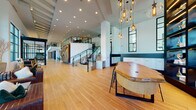 This expansive lobby and amenity space makes a strong architectural statement through its soaring double-height ceilings, warm wood-look flooring, and crisp white walls accented with bold navy detailing. At the heart of the space, a dramatic sculptural wood wall installation serves as a striking focal point alongside a wide staircase with sleek black metal railings leading to an open mezzanine level above.

Natural light pours in through oversized black-framed windows that frame views of the surrounding streetscape, lending the space an open, airy quality throughout. A curated collection of pendant lighting — including globe clusters and a statement ring chandelier — adds warmth and visual interest at varying heights.

The layout flows seamlessly between distinct zones: a lounge area with plush sectional seating and poufs, a co-working area with communal tables and patterned accent walls, and high-top dining tables positioned beside floor-to-ceiling windows. A glass-enclosed conference room provides a defined workspace within the open plan.

A dedicated mailroom area features white brick walls with a two-tone navy lower half and banks of individual mailboxes, complemented by tall interior clerestory windows. Throughout, the space balances contemporary design with curated artwork and organic textures, creating a sophisticated yet welcoming communal environment.