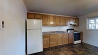 This single-floor space includes a kitchen designed for practical everyday use. The kitchen features an abundance of warm-toned wooden cabinetry, with both upper and lower cabinets providing generous storage capacity along the full length of the wall. Dark laminate countertops run continuously beneath the upper cabinets, offering ample prep space. The kitchen is equipped with a white refrigerator, a range with an electric stovetop, and an under-cabinet range hood for ventilation. A double-basin sink is centrally positioned along the counter with standard fixtures. The flooring consists of dark tile laid throughout, adding a grounded, neutral contrast to the lighter cabinetry. The walls are painted in a plain off-white tone, contributing to a clean, no-frills aesthetic. The upper cabinetry extends nearly to the ceiling, maximizing vertical storage space. Overall, the kitchen presents a straightforward, functional layout well-suited for everyday cooking and meal preparation, offering a practical and organized environment within this single-story space.
