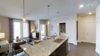 This single-story space showcases a thoughtfully designed open-concept layout that connects the kitchen, living, and bedroom areas in a cohesive, flowing arrangement. The kitchen serves as the focal point, anchored by a substantial island topped with speckled granite countertops and an undermount stainless steel sink. Dark cabinetry beneath the island provides a striking contrast to the light stone surface, while three pendant lights—featuring glass cylinder shades—hang overhead, offering both task lighting and a decorative touch.

The open layout allows clear sightlines from the kitchen island into the living area and through to the bedroom beyond, creating a sense of spaciousness throughout. The living area is furnished with comfortable seating and accented by a framed artwork piece, while large windows fitted with blinds and curtain panels allow natural light to filter in. The bedroom, visible in the background, continues the neutral color palette.

Dark hardwood-style flooring runs throughout, providing a grounding contrast to the light-toned walls and ceilings. Additional storage is accessible through white paneled doors, including what appears to be a pantry or utility closet. The overall ambiance is modern, clean, and well-appointed.