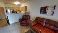 This single-floor space offers a practical and comfortable layout centered around an open-concept design that seamlessly connects the living area to the kitchen. The living area is furnished with rich burgundy leather seating, including a recliner and a loveseat, creating a warm and inviting atmosphere. Neutral-toned carpet runs throughout the living space, complementing the light gray walls. A decorative piece of wall art adds a pop of color and personality to the room. The kitchen is visible from the living area, separated by a low half-wall that functions as a breakfast bar or pass-through counter. It features warm honey-toned wood cabinetry, a stainless steel microwave, and a matching range, providing all the essentials for everyday cooking. Recessed and flush-mount ceiling lighting illuminates both areas efficiently. A baseboard heating unit along the living room wall ensures year-round comfort. The overall ambiance of the space is understated and functional, with a cozy, lived-in character that makes it well-suited for everyday residential use.