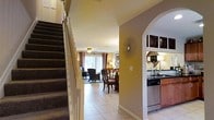 This two-story home offers a welcoming and functional layout designed for comfortable everyday living. The main level is anchored by an open-concept arrangement that seamlessly connects the living, dining, and kitchen areas. Light tile flooring flows throughout the main level, lending a clean and cohesive look to the space.

The living area is bathed in natural light streaming through large windows dressed with warm, decorative curtains, creating an inviting atmosphere. Nearby, the dining space comfortably accommodates a full dining set, positioned to take advantage of the open sightlines across the main floor.

Through a graceful archway, the kitchen is thoughtfully appointed with dark countertops, wood-toned cabinetry, a stainless steel sink, and a dishwasher — balancing practicality with style. A decorative starburst wall accent and a round mirror add personality and charm to the common areas.

A carpeted staircase with a classic white balustrade rises from the entry foyer to the upper level, offering a traditional architectural detail that anchors the home's design. The warm, golden wall tones throughout the main level contribute to the overall cozy and well-maintained ambiance of this multi-level home.