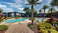 Located at 225 Sunlight Ln in Lake Mary, Florida, this outdoor amenity area offers a resort-inspired retreat defined by thoughtful design and abundant tropical landscaping. At its centerpiece sits a generously sized freeform swimming pool with crystal-clear water, surrounded by a paved brick deck outfitted with cushioned lounge chairs and sun loungers positioned to maximize relaxation and sun exposure. Mature palm trees, live oaks draped in Spanish moss, and vibrant tropical plantings of ferns, bromeliads, and ornamental shrubs frame the entire space, creating a lush, garden-like atmosphere. A standout feature is the pair of striking blue canopied cabanas with flowing curtains and circular built-in seating, providing a shaded, private retreat adjacent to the pool deck. A classic wood pergola anchors a secondary lounge area, offering additional covered outdoor seating amid carefully manicured garden beds. Herringbone and cobblestone brick pathways wind throughout the grounds, connecting the various amenity zones seamlessly. Iron fencing with integrated landscaping defines the perimeter, maintaining an open yet intimate feel. The multi-story residential buildings surrounding the space feature neutral gray and blue exteriors with balconies overlooking the amenity area, completing the cohesive community aesthetic.