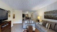 This single-floor residence features an open-concept design that seamlessly connects the living and dining areas, creating a cohesive and functional layout. The living room is anchored by two grey upholstered sofas arranged around a dark wood coffee table, accented with patterned throw pillows that add visual interest against the neutral color palette. Soft, wall-to-wall carpeting in a warm beige tone runs throughout the space, contributing to a comfortable underfoot feel. The smooth, light-grey ceiling with flush-mount lighting keeps the space feeling clean and uncluttered. Neutral white walls provide a versatile backdrop, complemented by decorative wall art and a tall indoor plant that introduces a natural element. Transitioning into the dining area, a round dining table with matching chairs sits beneath a ceiling light, framed by additional wall décor. A standard door provides access to an adjacent room or exterior. The overall ambiance is understated and practical, with a cohesive neutral scheme that lends itself well to a variety of furnishing styles.