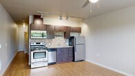 This efficient single-floor unit offers a thoughtfully designed layout that maximizes functionality within a compact footprint. The open-plan kitchen and living area form the heart of the space, featuring warm hardwood-style flooring that extends throughout. The kitchen is well-appointed with a full suite of stainless steel appliances, including a range with oven, over-the-range microwave, dishwasher, and refrigerator. Rich dark-stained cabinetry provides ample storage, while a mosaic tile backsplash in neutral tones adds a contemporary design accent. Track lighting overhead ensures excellent task illumination across the kitchen and living areas, complemented by a ceiling fan that enhances air circulation. The living area benefits from a large window with horizontal blinds, inviting natural light and offering pleasant outdoor views of surrounding greenery. The full bathroom continues the cohesive design aesthetic, featuring a dark wood vanity with a single sink, mosaic tile backsplash detail, tile flooring, and a full-size soaking tub with shower. A louvered closet door near the bathroom entry provides convenient storage access. The overall ambiance is clean, modern, and practical, making efficient use of every square foot.