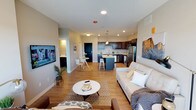 This single-floor residence is designed around an open-concept layout that integrates the living, dining, and kitchen areas into one cohesive, flowing space. The living area is anchored by a comfortable seating arrangement centered around a round coffee table, with warm hardwood-style flooring extending throughout. A large wall-mounted television is positioned near a hallway that leads to additional rooms, maintaining the open feel while providing a functional entertainment zone.

The kitchen is a standout feature, equipped with dark cabinetry, stainless steel appliances, and a central island with bar seating — ideal for casual dining or food preparation. Pendant lighting above the island adds a stylish accent while ensuring practical illumination. Recessed lighting throughout the main living space provides consistent, bright coverage.

Adjacent to the kitchen, a compact dining nook accommodates a small round table and chairs, offering a defined yet connected dining space. The neutral color palette of soft grays and warm whites, combined with natural light filtering through the space, creates an airy and welcoming ambiance. Overall, the layout maximizes functionality while maintaining a clean, contemporary aesthetic.