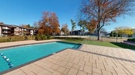 Located at 9007 Ten Mile Rd in Knoxville, Tennessee, this outdoor patio and amenity space offers a well-designed recreational environment set against a backdrop of mature trees and open skies. At the heart of the space is a generously sized swimming pool with clear turquoise water, framed by broad, light-toned paver decking that provides ample room for lounging and movement. The pool area is enclosed by a sleek black wrought iron fence, lending both security and a refined aesthetic to the setting.

Adjacent to the pool, a thoughtfully landscaped courtyard features ornamental trees set in circular stone-rimmed beds, trimmed shrubs, and lush green turf, creating a welcoming and well-maintained atmosphere. A pergola structure adds architectural character to the outdoor space, offering a shaded gathering spot.

The centerpiece building is a white-sided, multi-gabled structure with a dark shingled roof, large windows, and dark-trimmed double entry doors. Raised planting beds with neatly manicured shrubs line the building's exterior, enhancing its curb appeal. The surrounding grounds are enclosed by iron fencing with mature deciduous trees providing natural shade and seasonal color. The overall ambiance is clean, organized, and inviting, making this outdoor space a standout community amenity.