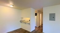 This single-floor space showcases a straightforward, functional layout designed to maximize efficiency. The living area flows seamlessly toward the kitchen zone, which is defined by a built-in pass-through counter with bracket supports and a granite-style surface — a practical feature that doubles as a casual dining or prep surface. The kitchen itself is equipped with a white electric coil cooktop integrated into the counter, a white refrigerator, and dark upper cabinetry, creating a simple contrast against the light walls. Throughout the space, wood-style plank flooring adds warmth and visual continuity, grounding the otherwise neutral palette of crisp white walls and ceilings. A hallway visible from the main living area leads toward additional rooms, likely including a bathroom, as suggested by the glimpse of white fixtures in the background. The layout feels open and uncluttered, with clean lines and minimal architectural ornamentation. The overall ambiance is bright and move-in ready, suited for those seeking a no-frills, practical living environment with an efficient use of space.