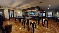 Located in State College, Pennsylvania, this single-floor entertainment space is designed as a sophisticated game room and lounge, combining recreational amenities with upscale interior finishes. The centerpiece of the room is a custom-built bar enclosure finished in dark beadboard paneling, topped with a rich granite-style countertop and equipped with upper cabinetry, display shelving, a mounted flat-screen TV, and bar-height seating along the front. Recessed lighting beneath a dropped soffit illuminates the bar area, adding a polished, venue-like atmosphere. The open floor plan accommodates a full-size pool table positioned beneath an industrial-style pendant light fixture, as well as a regulation shuffleboard table along one wall, flanked by wall-mounted décor and a flat-screen TV. High-top tables paired with black metal barstools are distributed throughout the space, providing flexible seating for guests. A tufted black leather booth-style banquette runs along one windowed wall, offering a comfortable lounge nook beneath wood-slatted blinds and decorative display shelving. The highly polished epoxy flooring in warm amber and brown tones adds a dramatic, reflective quality throughout the entire space, tying together the dark wood tones and neutral walls for a cohesive, upscale recreational aesthetic.