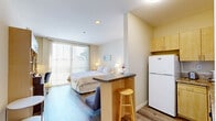 Located at 609 Broadway in the heart of Santa Monica, CA, this efficiently designed one-bedroom, one-bathroom unit offers a smart, open-concept layout that maximizes every square foot. The main living area seamlessly integrates the bedroom and living space, anchored by warm hardwood-style flooring throughout. A large sliding glass door floods the room with natural light and provides outdoor views, creating an airy, bright ambiance despite the compact footprint.

The bedroom area is generously proportioned relative to the overall unit size, accommodating a queen bed and featuring a built-in wall unit with ample cabinetry, shelving, a media center, and a dedicated workspace with a desk. A wall-mounted mini-split air conditioning unit ensures year-round comfort. A small breakfast bar with seating separates the living area from the kitchen zone.

The kitchen is fully equipped with light maple cabinetry, granite-style countertops, a full-size refrigerator, electric range, microwave, and ample storage — ideal for everyday cooking. A hallway leads to the bathroom, which features light tile flooring, a vanity with storage, and a clean, neutral palette. A closet provides additional storage. The unit's modern, minimalist design and prime Santa Monica location make it a highly functional and well-appointed space.