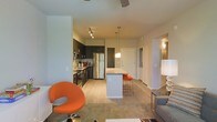 This single-floor residence offers a thoughtfully designed open-concept layout that integrates the living, dining, and kitchen areas into one cohesive and functional space. The kitchen is outfitted with dark cabinetry, stainless steel appliances including a refrigerator and microwave, and a sleek range. A central island with a light stone countertop serves as both a prep surface and casual dining spot, complete with orange bar stools that add a vibrant pop of color. Pendant lighting hangs above the island, providing focused illumination that complements the track lighting installed in the kitchen area. The living space features light wood-style flooring that transitions smoothly from the kitchen, while a plush area rug anchors the seating area. A bold orange accent chair adds a modern, eclectic touch to the living room. The overall color palette is neutral and contemporary, with warm white walls and ceilings that enhance the sense of openness. A hallway visible from the main living area leads to additional rooms, and a white entry door with a decorative wall sconce nearby provides a welcoming arrival point. The space blends modern design with functional living in a bright, open environment.