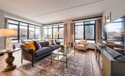 This single-floor residence offers a thoughtfully designed layout that balances open, communal spaces with well-appointed private retreats. The expansive living room serves as the heart of the home, featuring warm hardwood flooring, a gently angled ceiling, and a sweeping bank of large, black-framed windows that frame elevated urban cityscape views and flood the space with natural light. The open arrangement comfortably accommodates distinct seating and lounge areas, lending the room a generous, airy quality.

The kitchen is finished in a striking contrast of dark cabinetry and white subway tile backsplash, paired with stainless steel appliances and a light-toned countertop island that doubles as a casual dining space. Recessed lighting enhances the clean, modern aesthetic. An adjoining dining area benefits from additional window access and natural light.

The bedroom is serene and well-proportioned, featuring soft carpeting, a tall upholstered headboard wall, and large windows overlooking the city skyline. Two bathrooms offer distinct finishes: one features a walk-in shower fully clad in white subway tile with penny-round mosaic flooring and a glass enclosure, while the second includes a soaking tub with matching subway tile surround. Together, these spaces create a cohesive and refined urban residence.
