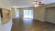 This single-floor space presents an open, combined living and dining area finished with rich, dark hardwood-style flooring that extends throughout. The layout is spacious and well-defined, with a clear visual separation between the dining area—illuminated by a classic pendant light fixture and framed by a bright window with blinds—and the adjoining living area, which features a ceiling fan with integrated lighting. The textured ceiling adds subtle architectural character to the space. Neutral, warm-toned walls complement the flooring and contribute to a cohesive, inviting atmosphere. A large sliding glass door serves as a prominent focal point, flooding the interior with natural light and offering views of a lush, tree-lined outdoor balcony. The balcony is accessible directly from the living area, creating a seamless indoor-outdoor connection. White baseboards provide clean contrast against the darker flooring, enhancing the overall finish of the space. The combination of natural light, open layout, and quality finishes makes this a functional and comfortable living environment.