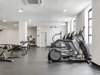 This fitness center offers a generously sized, single-floor workout facility designed to support a wide range of exercise routines. The space features a clean, modern aesthetic with crisp white walls, dark matte flooring, and recessed ceiling lighting that creates a bright, energizing atmosphere. Structural columns divide the floor plan into naturally flowing zones without compromising the overall sense of openness.

The cardio area is well-stocked with treadmills, elliptical trainers, stair climbers, stationary bikes, and a rowing machine, all positioned to take advantage of natural light from multiple large arched windows along the perimeter. A dedicated strength training zone houses a cable and functional trainer rig, along with a comprehensive free weights section featuring a full dumbbell rack, barbells, kettlebells, medicine balls, and adjustable benches.

An open stretching and functional training area provides ample floor space for bodyweight exercises, mobility work, and more. Additional amenities include a wall-mounted water fountain and hand sanitizer station. The facility is climate-controlled with ceiling-mounted HVAC units. The overall layout is thoughtfully organized, offering clear sightlines throughout and a welcoming, professional environment suitable for a full-body fitness routine.