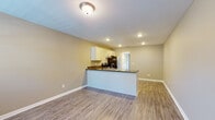 This single-floor unit offers a functional and cohesive layout centered around an open-concept living and kitchen area. Wood-look luxury vinyl flooring flows consistently throughout, complementing the neutral warm-beige walls and crisp white baseboards to create a clean, unified aesthetic.

The kitchen serves as a focal point of the space, featuring a peninsula with dark granite-style countertops that provides both workspace and a natural divider between the kitchen and living area. White shaker-style cabinetry with brushed hardware offers generous storage, while stainless steel appliances — including a refrigerator, range, and dishwasher — complete the functional setup. Recessed lighting illuminates the kitchen efficiently.

The open living area is spacious and adaptable, receiving natural light from a large sliding glass door fitted with vertical blinds. The door opens to a view of mature trees, adding a pleasant outdoor connection to the interior.

The bathroom is finished with a full-sized tub-shower combination, white surround, and wood-look flooring consistent with the rest of the unit. Overall, the space presents a straightforward, well-maintained design with practical finishes throughout.