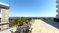 This exceptional rooftop terrace is a standout outdoor retreat, designed to maximize its breathtaking surroundings. The expansive stone tile floor is laid in a clean, large-format pattern that lends a polished, refined aesthetic to the space. A sleek wrought iron railing borders the perimeter, ensuring unobstructed sightlines while providing a sense of security at elevation.

The terrace is furnished with classic wrought iron tables and chairs, thoughtfully arranged to take full advantage of the panoramic views. Potted plants add touches of greenery, softening the hardscape and bringing warmth to the open-air environment. Lounge chairs are positioned to invite relaxation under the open sky.

The views from this terrace are truly remarkable — a vast, uninterrupted expanse of open water stretches to the horizon, framed by a canopy of mature trees below. The elevated vantage point provides a serene and private atmosphere, well above the surrounding landscape.

Flooded with natural light and open to clear blue skies, this terrace offers an ideal setting for outdoor entertaining, dining, or simply unwinding while taking in the sweeping waterfront scenery. It is a rare and distinguished outdoor space that combines elegant design with an extraordinary natural backdrop.