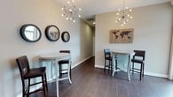 Located in Jefferson, this single-floor space showcases a clean, contemporary design with a welcoming and polished ambiance. The interior is defined by large-format dark tile flooring that runs throughout, providing a sleek contrast to the neutral, light-toned walls. The space is illuminated by two striking sputnik-style chandeliers, each featuring starburst arms with exposed globe bulbs that serve as both functional lighting and bold decorative focal points. Seating is arranged around bistro-style high-top tables paired with dark upholstered bar stools, creating an informal yet sophisticated social atmosphere well-suited for gathering or casual meetings. Wall décor includes a series of round dark-framed mirrors and a rustic wood-panel world map artwork, adding layers of texture and visual interest to the space. A second world map wall piece with an inspirational quote reinforces the travel-inspired aesthetic theme carried throughout. The layout is open and airy, with a corridor visible in the background suggesting additional areas beyond. The combination of modern lighting, coordinated furniture, and thoughtfully curated décor gives the space a refined yet approachable character that is both functional and visually engaging.