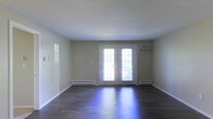 This single-floor space offers a clean and functional layout with a neutral, understated aesthetic. The main living area is characterized by rich, dark hardwood flooring that adds warmth and contrast to the light-toned walls and white trim throughout. A standout feature of the room is the set of French doors centered on the rear wall, allowing an abundance of natural light to pour in while providing a view of the outdoors. Flanking the French doors are windows dressed with blinds, further enhancing the brightness of the space. Baseboard heaters run along the base of the walls, ensuring efficient heating, while a wall-mounted air conditioning unit positioned near the French doors provides climate control during warmer months. The smooth, white ceiling and clean wall surfaces give the interior a fresh and open feel, making it a versatile canvas ready to accommodate a variety of uses or furnishing styles. The overall ambiance is bright, practical, and well-maintained, suited for those seeking a straightforward, move-in-ready space.