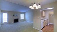 This single-floor residence offers a comfortable and functional layout designed for everyday living. The main living area is a standout feature, showcasing soft wall-to-wall carpeting underfoot and a classic fireplace with a clean white mantel that serves as a natural focal point. Vaulted ceilings lend a sense of openness and airiness to the space, while a window allows natural light to fill the room with a warm, inviting glow. An elegant multi-arm chandelier hangs prominently, adding a decorative touch that bridges classic and casual design sensibilities. The living space flows seamlessly into the kitchen, which is accessible through an open passageway. The kitchen features white cabinetry, a sink positioned beneath additional upper cabinets, and light hardwood-style flooring that contrasts nicely with the carpeted living area. The overall color palette throughout the home is soft and neutral, with light gray-green walls and crisp white trim creating a clean, cohesive aesthetic. The open-concept connection between the main living and kitchen areas promotes easy movement and interaction, making the space well-suited for both relaxed daily living and casual entertaining.