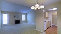 This single-floor residence offers a comfortable and functional layout designed for everyday living. The main living area is a standout feature, showcasing soft wall-to-wall carpeting underfoot and a classic fireplace with a clean white mantel that serves as a natural focal point. Vaulted ceilings lend a sense of openness and airiness to the space, while a window allows natural light to fill the room with a warm, inviting glow. An elegant multi-arm chandelier hangs prominently, adding a decorative touch that bridges classic and casual design sensibilities. The living space flows seamlessly into the kitchen, which is accessible through an open passageway. The kitchen features white cabinetry, a sink positioned beneath additional upper cabinets, and light hardwood-style flooring that contrasts nicely with the carpeted living area. The overall color palette throughout the home is soft and neutral, with light gray-green walls and crisp white trim creating a clean, cohesive aesthetic. The open-concept connection between the main living and kitchen areas promotes easy movement and interaction, making the space well-suited for both relaxed daily living and casual entertaining.