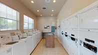 This shared laundry facility is designed for convenience and efficiency, offering a clean and organized environment for residents. The room is laid out with a row of top-load washing machines along one wall, providing ample capacity for laundry needs. Opposite, a bank of stacked dryer units maximizes the available space, offering multiple drying options simultaneously. At the center of the room stands a freestanding island unit with a built-in utility sink, offering a practical space for pre-treating garments or hand-washing delicate items. The light tile flooring is easy to maintain and contributes to the room's bright, tidy appearance. Recessed ceiling lights illuminate the space evenly, complemented by natural light filtering in through a window fitted with blinds. A bulletin board mounted near the entrance provides a space for community notices and guidelines, while clearly posted instructions ensure ease of use for all. A fire extinguisher is also present, reflecting attention to safety standards. The neutral, warm-toned walls and clean white appliances give the facility a welcoming and functional atmosphere suited to everyday use.
