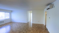 This single-floor space offers a straightforward and functional layout characterized by its classic parquet wood flooring, which adds a timeless warmth throughout the interior. The open main living area is finished with crisp white walls and a smooth ceiling, creating a bright and airy atmosphere that reflects natural light effectively. The parquet flooring, laid in a traditional basket-weave pattern, is a standout architectural detail that lends character to the space. A central hallway connects the main living area to additional rooms beyond, providing a logical and practical flow through the layout. Doorways are cleanly framed and allow for easy transition between spaces. Baseboard heating units are present along the walls, indicating a reliable in-unit heating system. The neutral color palette throughout keeps the space feeling open and versatile, making it easy to furnish and personalize. Overall, the space presents a clean, well-maintained interior with classic design elements and a functional, connected floor plan suited for comfortable everyday living.