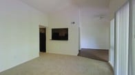 This single-story space showcases an open-concept layout defined by dramatically vaulted ceilings that lend a sense of spaciousness and airiness to the main living area. The living room is finished with plush carpeting and anchored beneath a ceiling fan, which aids in air circulation throughout the generous vertical space above.

A defining architectural feature is the pass-through window connecting the kitchen to the living area, offering both visual openness and functional convenience. The kitchen, visible through the pass-through and an adjacent doorway, features dark cabinetry and includes a refrigerator and range, providing a contrast to the lighter tones of the surrounding walls.

The transition from carpeted living space to hard flooring marks a natural separation between areas, adding subtle definition to the open floor plan. The walls throughout are finished in a neutral, light tone that reflects available light and enhances the bright, clean atmosphere of the interior. A ceiling-mounted HVAC vent is visible near the top of the vaulted wall, ensuring efficient climate control throughout the space. Overall, the layout balances functionality with an expansive, airy ambiance.