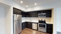 The kitchen is a standout feature of this single-floor space, designed with a modern aesthetic that balances functionality and style. Dark espresso-toned cabinetry lines the walls from base to upper level, providing generous storage while creating a bold visual contrast against the crisp white countertops and bright white subway tile backsplash. The full suite of stainless steel appliances includes a refrigerator, electric range with oven, built-in microwave, and dishwasher, offering a cohesive and polished look throughout. The L-shaped layout makes efficient use of the available space, providing ample countertop workspace for meal preparation. A double-basin undermount sink is positioned beneath upper cabinetry, maintaining a clean sightline throughout the room. Recessed lighting in the white ceiling ensures the kitchen is well-lit, enhancing the overall brightness of the space. The warm-toned hardwood flooring grounds the design, adding natural texture that softens the contrast between the dark cabinetry and light surfaces. The overall ambiance is contemporary and refined.
