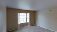This single-floor space features a spacious, open room finished with wall-to-wall carpet in a neutral beige tone, providing a soft and comfortable underfoot feel. The room is defined by its standout bay window, which projects outward from the wall and is fitted with horizontal blinds, allowing for adjustable natural light control. The multi-panel bay window configuration creates a pleasant architectural focal point while drawing in diffused outdoor light, contributing to an airy, open atmosphere. The walls are painted in a warm, neutral beige that complements the light carpeting, creating a cohesive and understated color palette throughout the room. White baseboards run along the perimeter, adding a crisp contrast to the warmer wall tones. The ceiling is smooth and painted white, enhancing the sense of openness in the room. The overall layout is straightforward and versatile, offering ample open floor space that lends itself well to a variety of furniture arrangements. The space presents a clean, move-in ready condition with a calm, neutral ambiance well-suited to a range of functional uses.