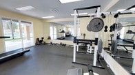 This dedicated fitness space is thoughtfully designed to support a full range of workout routines. The room is finished with durable black rubber flooring, providing both safety and sound absorption during exercise. Pale yellow walls lend a neutral, energizing atmosphere, while recessed fluorescent lighting and a skylight ensure the space is well-lit throughout the day. Windows along one wall allow natural light to filter in, enhancing the overall brightness of the room.

A large wall-mounted mirror spans a significant portion of one wall, visually expanding the space and allowing users to monitor their form during workouts. The fitness area is equipped with a heavy-duty power rack loaded with Olympic weight plates, a flat bench, and an assortment of dumbbells neatly arranged on a weight tree. Additional free weights and barbells are accessible on the floor nearby. Cardio equipment, including a treadmill and a stationary bike, is positioned toward the back of the room. A door provides convenient access to an adjacent exterior area, adding to the functionality of this compact yet comprehensively outfitted fitness space.