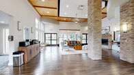 The Silverado leasing office and clubhouse is a thoughtfully designed single-floor facility that blends rustic warmth with modern functionality. The open-concept interior is immediately striking, featuring soaring ceilings accented with rich wood tongue-and-groove paneling and recessed lighting, creating an inviting, lodge-inspired atmosphere throughout.

Wood-look tile flooring runs seamlessly across the expansive main area, anchored by a prominent stacked-stone column that serves as a dramatic architectural centerpiece. A matching stacked-stone fireplace with a natural wood mantle anchors the lounge area, which is furnished with comfortable seating arranged around a warm coffee table — ideal for casual gatherings or relaxed conversations.

The leasing office area is clearly defined by carpeted flooring and a stacked-stone accent wall bearing the Silverado branding. Workstations with flat-screen monitors and ample seating accommodate both staff and visitors efficiently. A beverage station with cabinetry provides added convenience near the entry.

A dedicated dining area offers a large rustic wood table surrounded by upholstered chairs, bathed in natural light from floor-to-ceiling sliding glass doors that open to views of the resort-style pool and surrounding community. This seamless indoor-outdoor connection, paired with the cohesive use of natural stone, wood tones, and neutral walls, gives the entire space a welcoming and polished character.