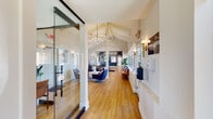 This single-floor amenity space makes a strong first impression with its welcoming entryway, where warm hardwood flooring and soaring vaulted ceilings with exposed white beam detailing set an inviting tone. A striking sputnik-style chandelier with brass accents anchors the space overhead, casting a warm glow across the open layout.

The main lounge area is thoughtfully furnished with contemporary navy blue armchairs arranged around a round marble-top coffee table, creating an ideal setting for socializing or relaxing. Glass partitions add transparency while defining zones within the space, and curated wall art adds personality throughout.

A built-in kitchenette serves as a functional focal point in the lounge, featuring deep blue subway tile backsplash, sleek cabinetry, a compact refrigerator, microwave, and countertop workspace — all accented by gold pendant lighting for a polished, modern touch.

The fitness center occupies a generously sized room distinguished by its dramatic vaulted ceiling with white-painted exposed rafters and large pendant lights. The space is well-appointed with cardio equipment including a treadmill and elliptical, a cable resistance machine, an adjustable weight bench, a full rack of dumbbells and kettlebells, and an array of exercise balls and functional training accessories. A wall-mounted flat-screen TV and natural light from surrounding windows complete the workout environment.